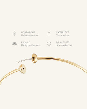 Two elegant Icon Hoop Earrings Medium by JENNYBIRD in high-polish gold are displayed against a light background. Each earring features a small spherical closure. One lies flat while the other stands upright, highlighting their minimalistic design crafted from stainless steel.