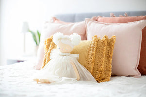 A young girl with light hair tied in a bow sits on a bed with white linens, smiling and holding up an Alimrose Ava Angel Doll Ivory Gold. She wears a light-colored dress, and behind her are pink and rust-colored pillows against a gray headboard.