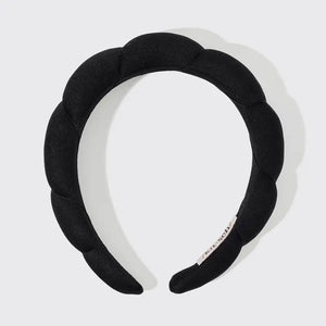 A person with long blonde hair is smiling and looking to the side. They are wearing the KITSCH PUFFY HEADBAND, a black padded accessory crafted from recycled polyester. The plain white background accentuates their cheerful expression and the eco-friendly headband.