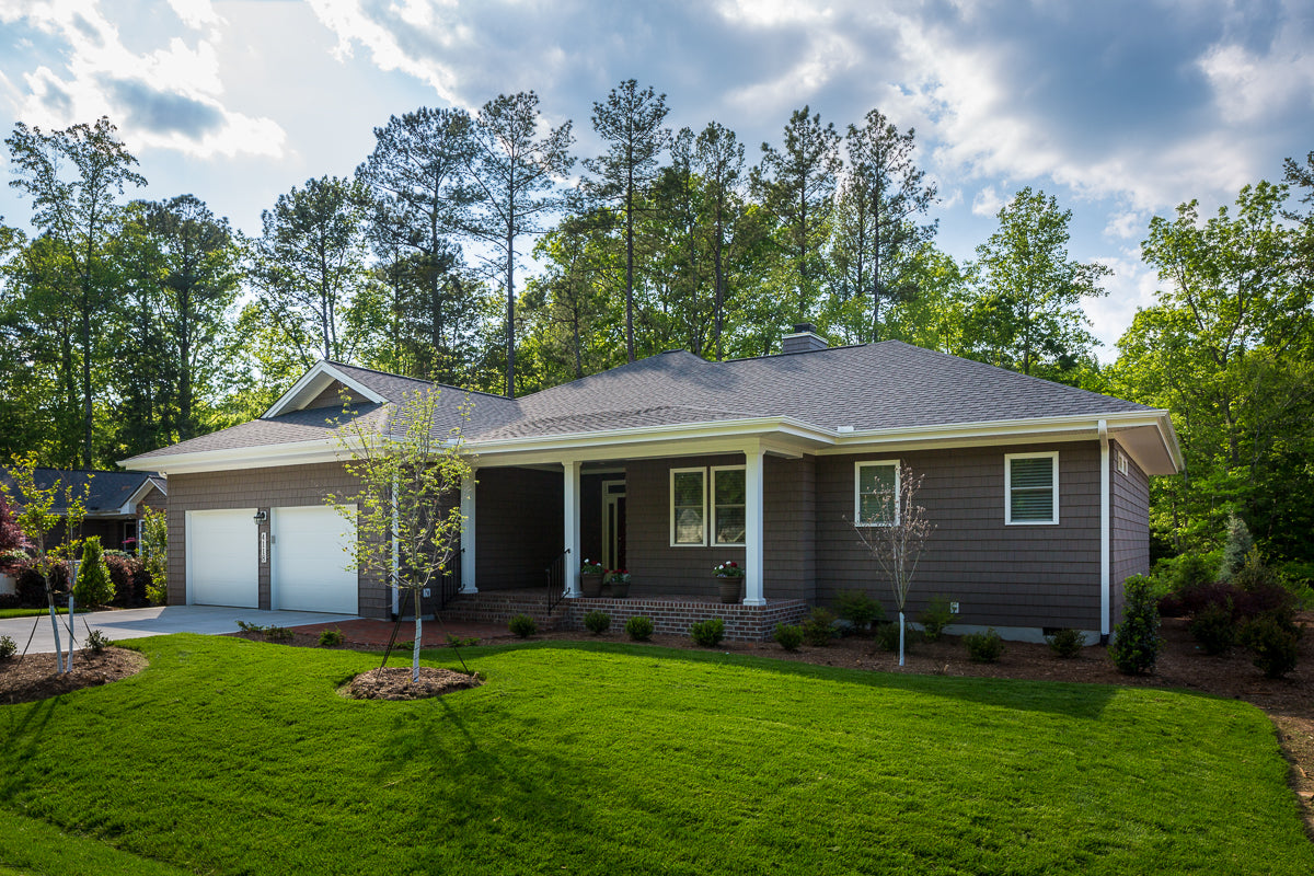 Single-story house with gray siding, a covered front porch, attached two-car garage, well-maintained lawn, and small landscaped trees, set against a backdrop of tall green trees under a partly cloudy sky.