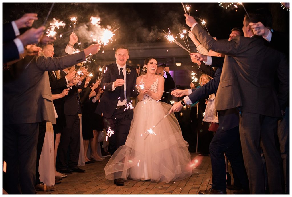 A bride and groom walk joyfully through a crowd of guests holding sparklers at night. The couple smiles and laughs, surrounded by well-dressed friends celebrating their wedding.