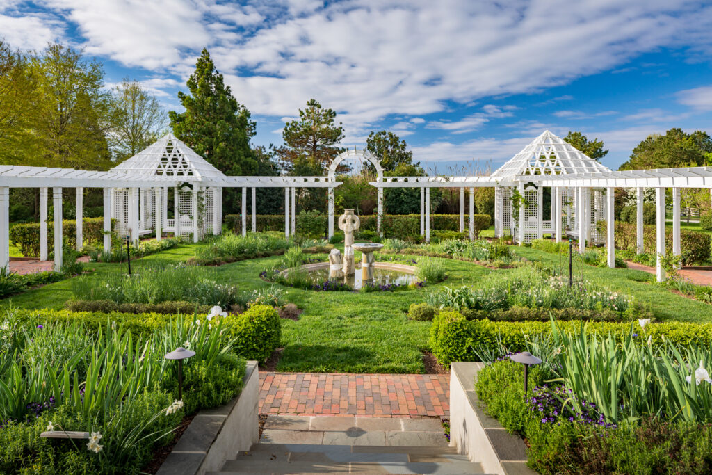 A formal garden features lush green lawns, blooming flower beds, a central fountain with a statue, white pergolas, and two white gazebo structures under a blue sky with scattered clouds.