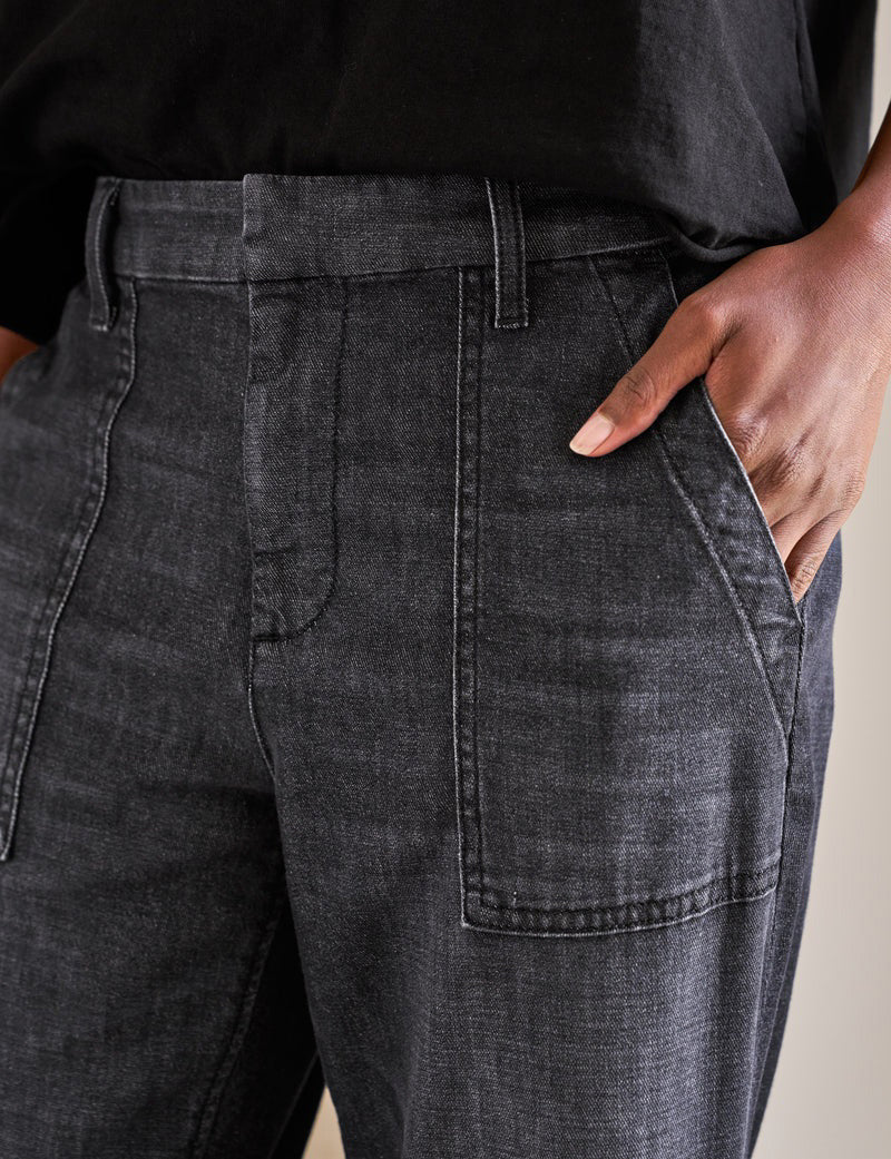 A person stands indoors with hands in the utility pockets of FRANK &amp; EILEEN’s Jameson Jogger Utility Jean in 1981 Wash. They pair it with a black top and tan ballet flats with black toe caps. A dining table and chairs are visible behind them.