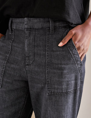 A person stands indoors with hands in the utility pockets of FRANK & EILEEN’s Jameson Jogger Utility Jean in 1981 Wash. They pair it with a black top and tan ballet flats with black toe caps. A dining table and chairs are visible behind them.