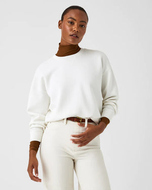 A woman stands confidently against a plain white background, wearing the SPANX Aireessentials Crew Neck Pullover and matching sweatpants made from luxurious comfort spacer fabric. She has her hair pulled back and her left hand is raised slightly. She smiles gently with her lips closed.