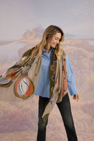 A woman with long blonde hair smiles at the camera in a scenic setting, wearing a white shirt, plaid pants, and the STORIATIPIC - ASTER WOOL SCARF by STORIATIPIC. Mountains, grass, and water are visible in the background.