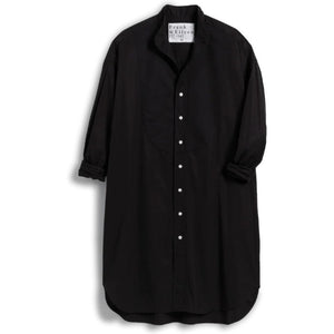 Dressed in the elegant FRANK & EILEEN TORI Italian Tuxedo Shirtdress in a sleek black hue, an individual casually stands with one hand tucked into the dress pocket and a glass of red wine held in the other. The scene is set indoors, radiating warmth with its cozy, furnished ambiance.