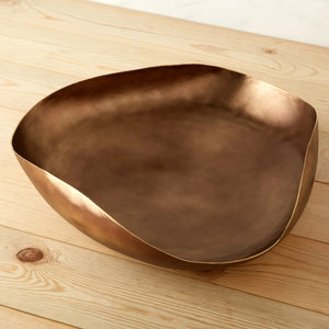 A round, deep bowl from BE HOME, INC's AGED BRONZE SERVING SET with a smooth, golden-bronze finish sits on a light wooden surface. The slightly irregular, organic shape of the bowl enhances its handcrafted appearance. The background displays the light wood grain pattern of the tabletop.