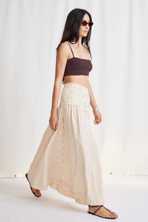 A woman with long hair is wearing sunglasses, a dark crop top, and the APIECE APART - ORA SMOCK MAXI SKIRT made of organic cotton. She is standing against a light-colored background and wearing sandals.