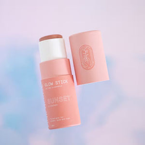 A cylindrical pink glow stick makeup product, with its cap off, is displayed against a blurred pastel pink and blue background. The label reads "PINK HOUSE ORGANICS - GLOW STICK SUNSET," indicating it’s suitable for lips, cheeks, and eyes to achieve a natural glow. The cap features a printed design.