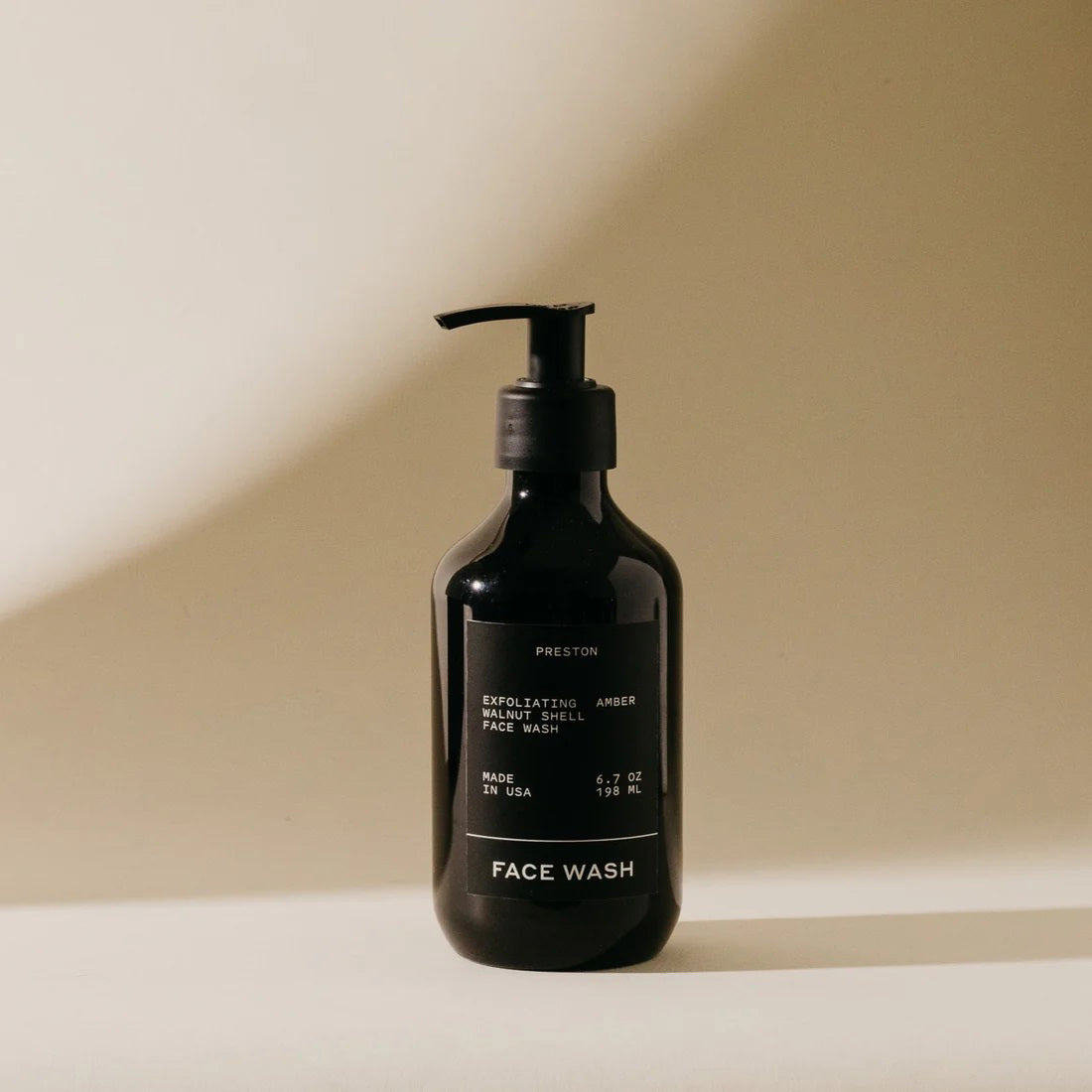 A black pump bottle labeled &quot;PRESTON - FACE WASH&quot; stands on a soft, neutral-toned surface. The bottle features a sleek, minimalist design with white text and exfoliating walnut shells. The light casts a shadow on the background, creating a calming atmosphere.