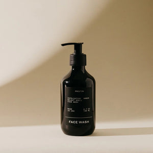 A black pump bottle labeled "PRESTON - FACE WASH" stands on a soft, neutral-toned surface. The bottle features a sleek, minimalist design with white text and exfoliating walnut shells. The light casts a shadow on the background, creating a calming atmosphere.