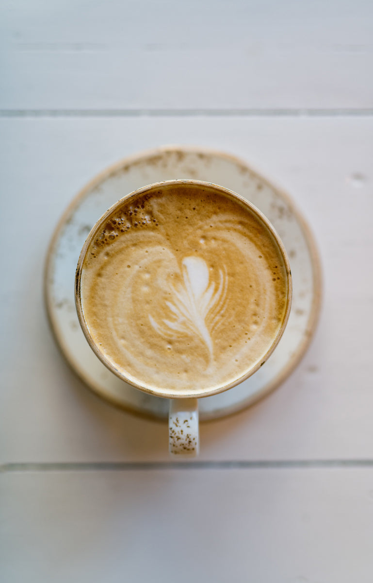 A cup of latte with delicate leaf-shaped latte art on top, placed on a speckled saucer on a white wooden surface, viewed from above.