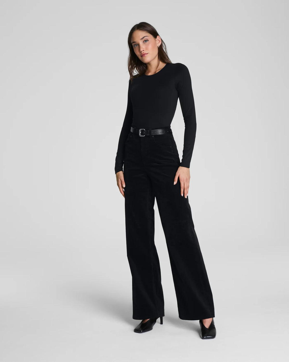 A person wears the SPANX Corduroy Wide Leg Jean in Black with a long-sleeve black top and black pointed-toe heels, standing against a plain light background.