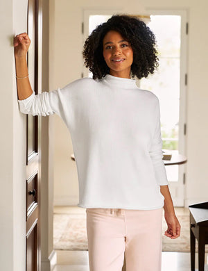 A woman with curly hair leans against a wall, smiling softly. She wears a white FRANK & EILEEN Monterey Rolled Funnel Neck Cotton Sweater and rests one hand near her chin. A blurred plant is in the background.