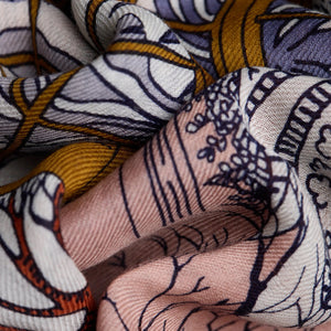 A close-up image of the INOUI EDITIONS - QUATRE SAISONS SCARF by INOUI, showcasing a lightweight summer accessory adorned with intricate, colorful patterns. The design features an array of floral and geometric shapes in shades of white, gold, and blue on a pinkish background, highlighting the cloth's textured surface and detailed print.