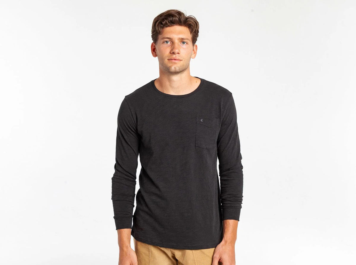 A young man with short brown hair wears the CRIQUET Cotton Slub Long Sleeve T-Shirt in black, featuring a chest pocket, paired with tan pants, standing against a plain white background.