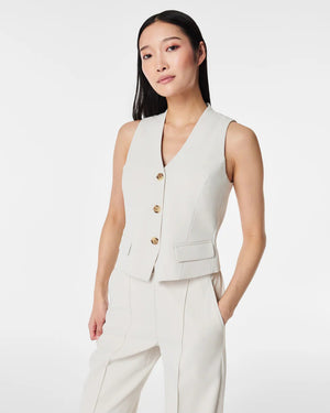 A person with long, straight hair is wearing the SPANX CAREFREE CREPE VEST, a light beige, sleeveless, tailored vest made of easy-care crepe fabric with three buttons and matching trousers from SPANX. Their hands are in their pockets as they pose in front of a plain white background.