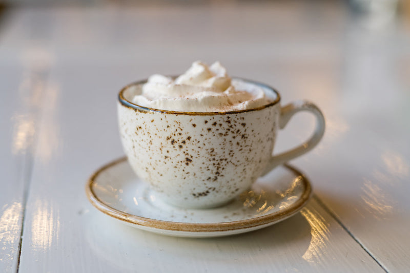 A speckled ceramic cup filled with a hot beverage topped with whipped cream, placed on a matching saucer on a shiny white table.