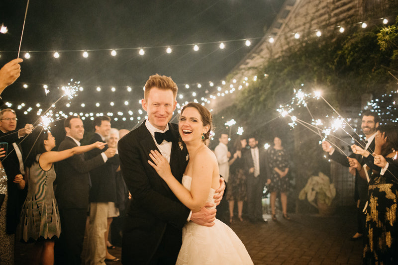 A bride and groom smile and embrace under string lights at night, surrounded by guests holding sparklers, creating a festive and joyful wedding celebration atmosphere.