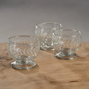 The HANDMADE ETCHED TEQUILA GLASS by ZODAX is a clear glass dessert bowl with a short stem and round base, measuring 3 inches by 2.75 inches. It features an etched leaf pattern around its exterior. The transparent glass reflects light, giving it a shiny appearance. The overall design is simple and elegant.