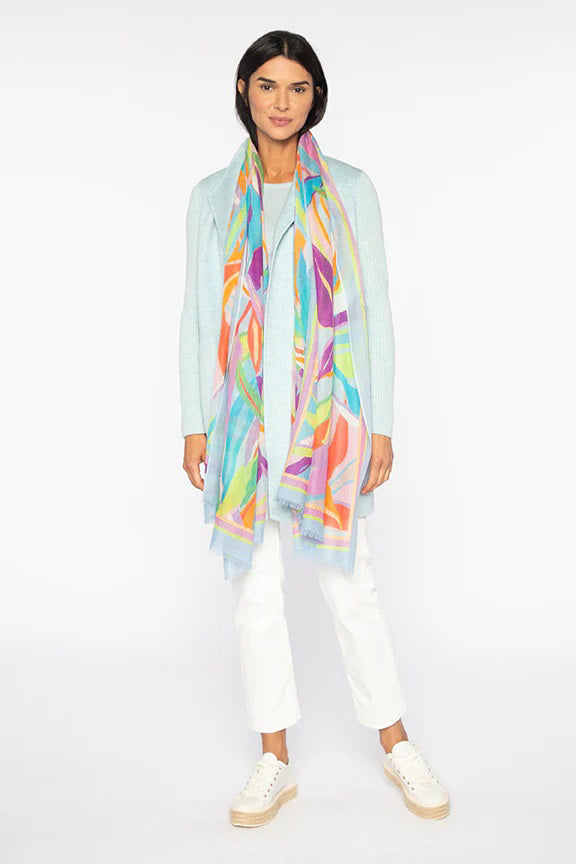 A person with long hair elegantly wears a light blue sweater, white pants, and sleek white shoes. Gracefully draped around their neck is the palm print scarf from KINROSS CASHMERE by KINROSS against a plain white background.