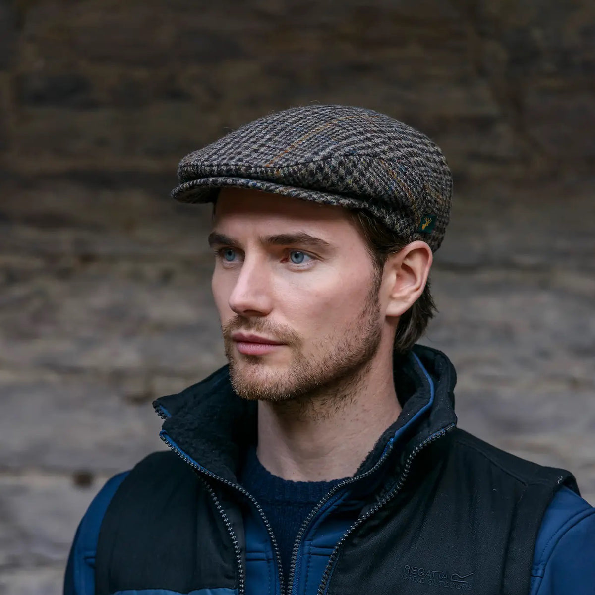 A man with light skin and short brown hair wears the MUCROS WEAVERS - TRINITY CAP by MUCROS WEAVERS, along with a navy jacket and black vest, standing before a stone wall and looking slightly to the right with a neutral expression.
