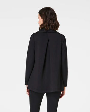 A person wearing the SPANX - AIRESSENTIALS DRAPE FRONT CARDIGAN in black, designed with long sleeves and crafted from lightweight fabric, elegantly poses with their hand in one of its pockets. The cardigan features an asymmetrical hem and is paired beautifully with black pants, highlighting the side and pocket details against a plain background.