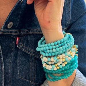 A person wearing a denim jacket showcases a stack of bracelets on their wrist, with the CC AND CO BY CATHERINE CANINO - SALLY BRACELET IN TURQUOISE standing out. Its stretchy fit bands and turquoise, white, and gold beads add natural color variations and texture to the accessory.