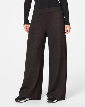 A person is wearing the SPANX AIRESSENTIALS WIDE LEG SHIMMER PANT in black along with a zip-up top. They stand casually with their hair tied back, sporting black and white sneakers, all set against a plain white background.