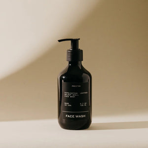 A black pump bottle labeled "PRESTON - FACE WASH" stands on a soft, neutral-toned surface. The bottle features a sleek, minimalist design with white text and exfoliating walnut shells. The light casts a shadow on the background, creating a calming atmosphere.