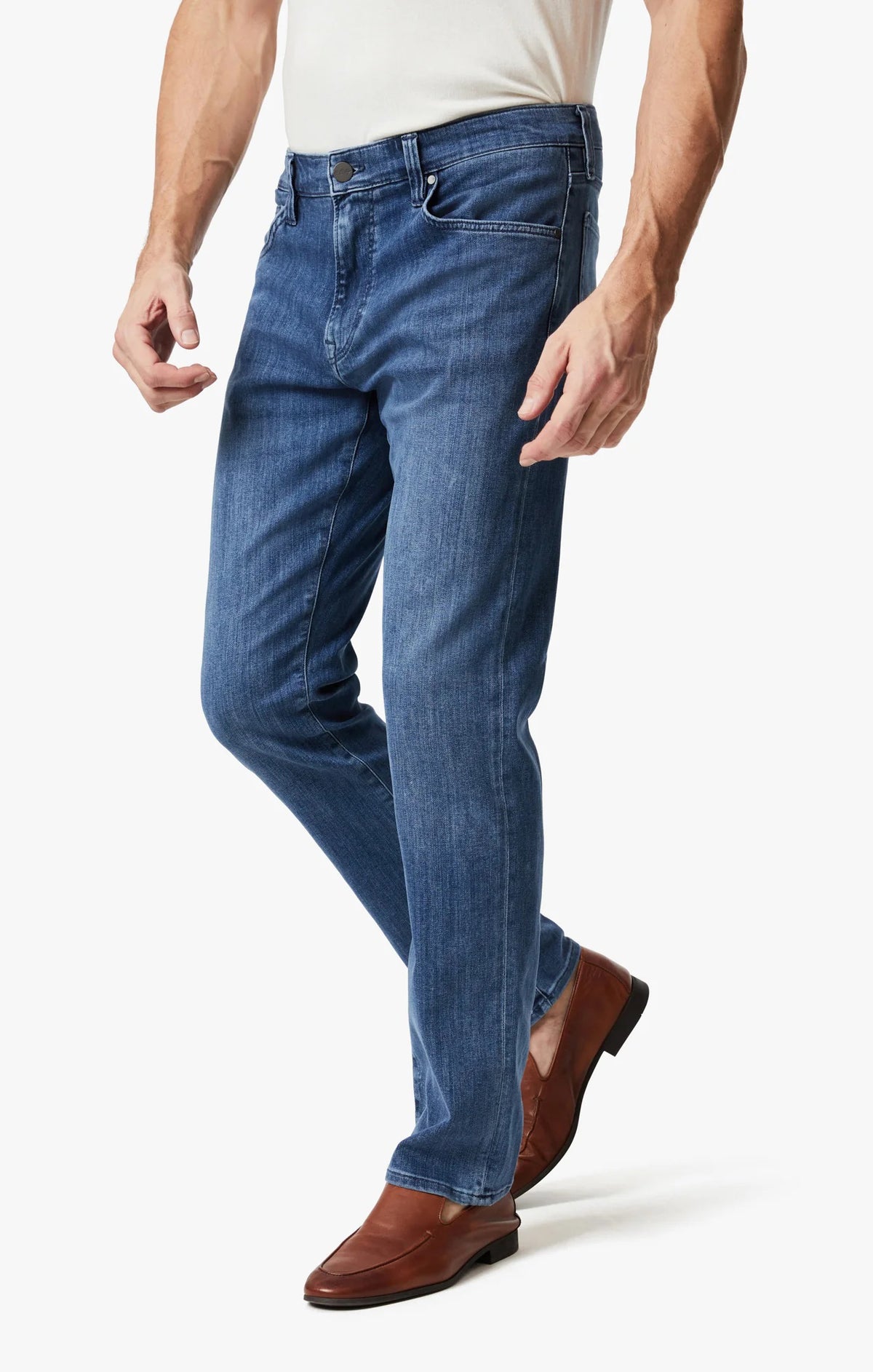 A man wears 34 HERITAGE CHARISMA Relaxed Straight Leg Denim in Mid Urban Wash, a white shirt, and brown leather shoes, standing against a plain white background.
