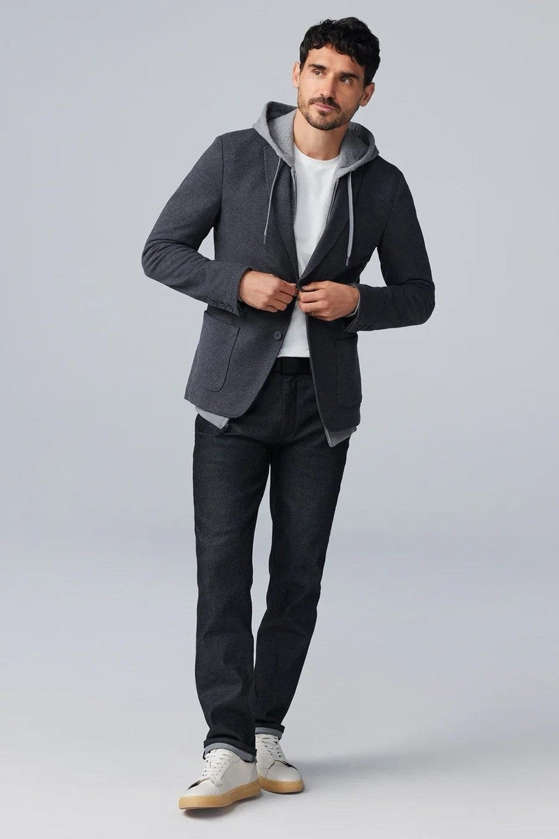 A man fastens the MACK WELDON Ace Blazer in Charcoal Heather over a gray hoodie, white tee, and dark jeans, paired with white sneakers with beige soles. He stands against a plain background, looking slightly to the side.