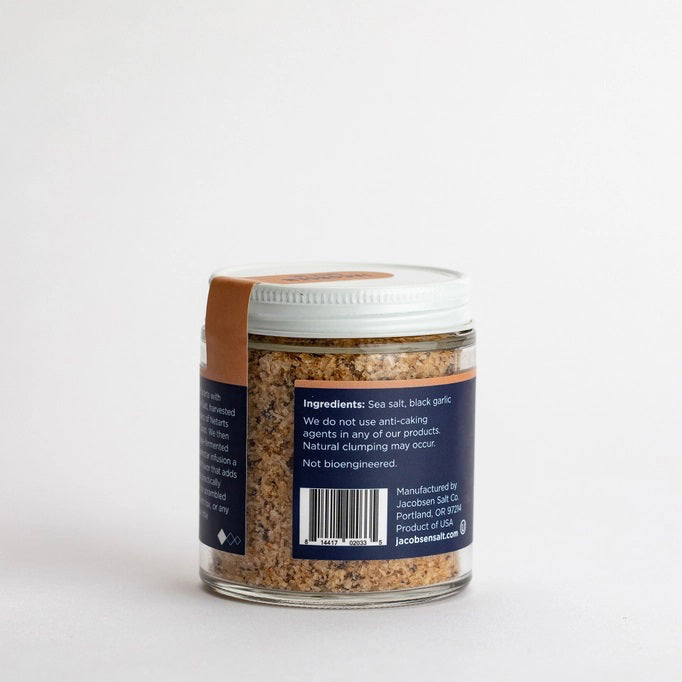 A small glass jar with a white lid, labeled "Jacobsen Salt Co. - Infused Black Garlic Salt," is placed on a light beige surface against a plain, light gray background. The jar contains an aromatic blend of coarse, brownish salt with rich garlic flavor.