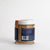 A small glass jar with a white lid, labeled "Jacobsen Salt Co. - Infused Black Garlic Salt," is placed on a light beige surface against a plain, light gray background. The jar contains an aromatic blend of coarse, brownish salt with rich garlic flavor.