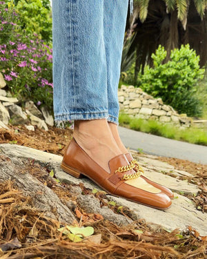 The FLAT TWO TONE LOAFER WITH CHAIN by MADISON MAISON is a stylish flat loafer crafted from the finest leathers. It features a light beige vamp adorned with two gold chain embellishments on the upper. Designed for maximum comfort, this brown leather beauty boasts a classic slip-on design and a low heel.