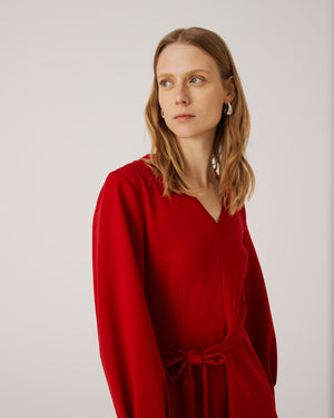 A woman with long, light brown hair stands against a plain background. She is wearing the SFIZIO long sleeve v-neck flare skirt dress, featuring a tie waist. Looking to her left with a calm expression, she has hoop earrings and exudes minimalist style.