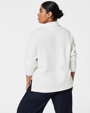 A person with dark hair styled in a neat updo is wearing the SPANX - AirEssentials Split Neck Polo Top in black, paired with matching black pants. They rest one hand on the back of their neck and gaze to the side against a plain white background, exuding an air of luxurious comfort.