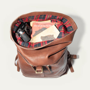 The WILL LEATHER GOODS - ROLL TOP BACKPACK by WILL LEATHER GOODS is open, showing a camera, a red-and-black plaid lining, an Andrzej Sapkowski book, and an old yellowed map inside.