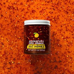 A jar of MOMOFUKU - CHILI CRUNCH HOT HONEY, with a white lid, contains a red-brown mixture inside. The label features an illustration of a citrus fruit along with the text "Momofuku Chili Crunch Hot Honey" and "Net Wt 7.7 oz (220g)." This spicy-crunchy chili oil offers the perfect blend of heat and sweetness.