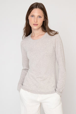 A person with long, brown hair is standing against a plain, light-colored background. They are wearing a KINROSS Cashmere - Long Sleeve Silk and Cashmere Crew in light gray and white pants. Their hands are in their pockets, and they are looking directly at the camera.