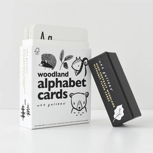 A set of WEE GALLERY - WOODLAND ALPHABET CARDS from Wee Gallery is displayed in an open white box, showcasing high-contrast illustrations of woodland animals and letters. Next to the box on a white surface lies a separate black card holder with the brand name "WEE GALLERY" and a leaf design, enhancing your child's learning experience.