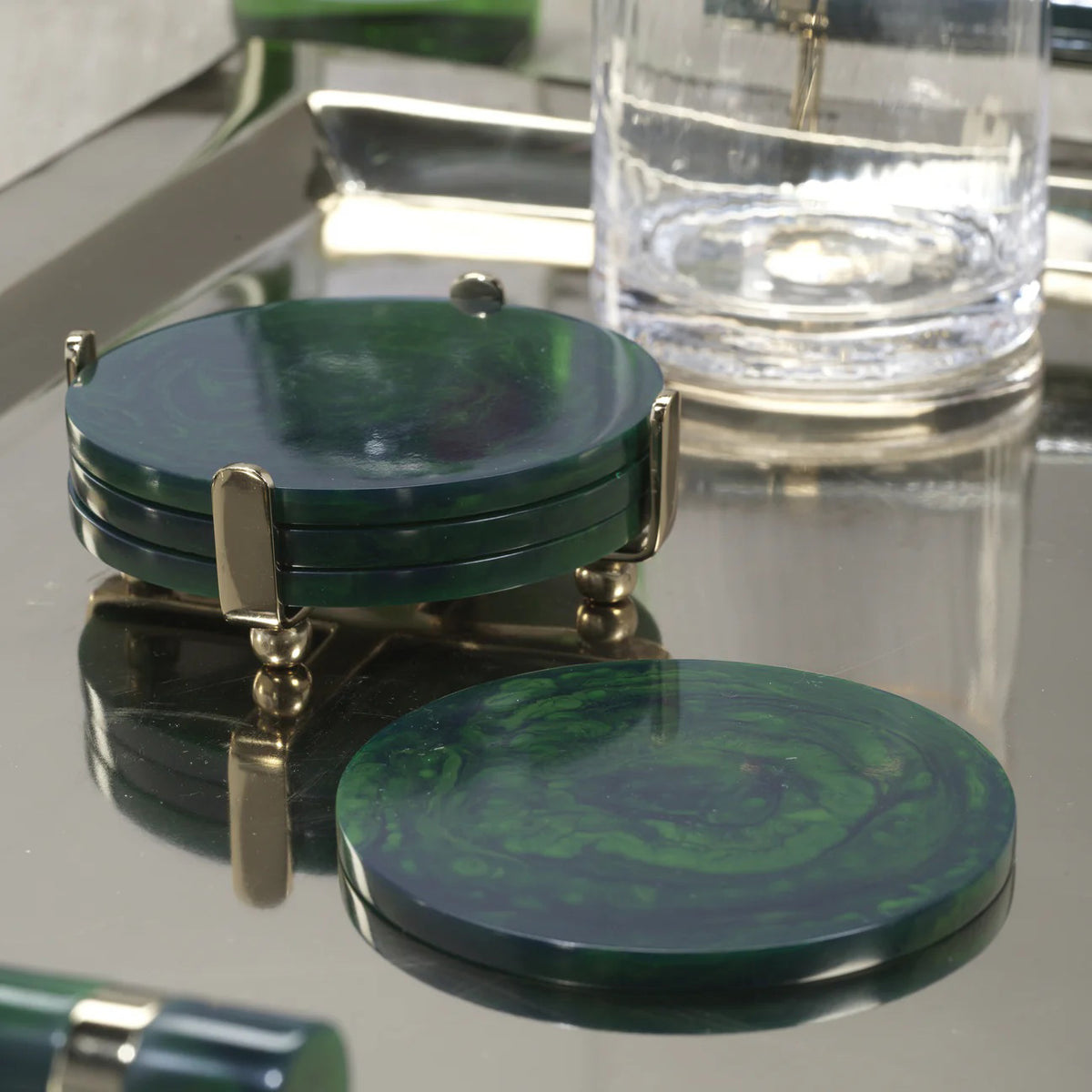 The ZODAX SPIGA Set of 4 round, dark green marbled resin coasters includes a metal stand with four posts. One coaster is displayed separately; the others are stacked in the holder. Size: 4.25” x 1.75”.
