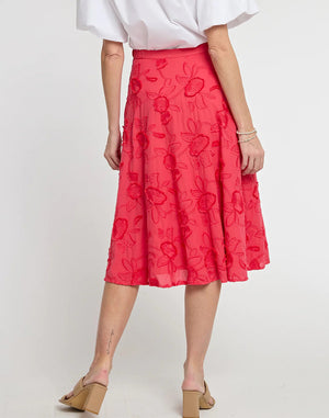 A person with short hair wears a white button-up shirt with puff sleeves and a high-waisted, knee-length Gloria Coral Floral Applique Skirt by HINSON WU. They also have beige heeled sandals and a bracelet, posing against a plain white background.