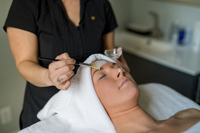 A person with a towel wrapped around their head lies on a spa bed while an esthetician applies a facial mask with a brush. The setting appears calm and relaxing.