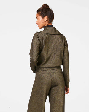 Dressed in the SPANX - AIRESSENTIALS HALF ZIP SHIMMER PULLOVER, a shimmering, gold-toned tracksuit crafted from ultra-soft fabric featuring a quarter-zip top and coordinating pants, a person stands against a simple white background with their hair styled neatly in a bun, radiating a gentle smile.