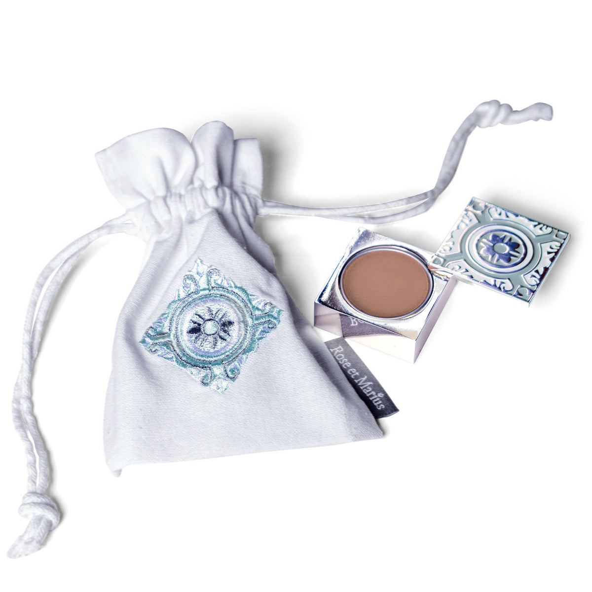 A manicured hand holds an open ROSE ET MARIUS &quot;A Stroll Under the Oratory&quot; solid perfume compact with a mirror and round perfume inside, evoking Haute Parfumerie, set against a white background.