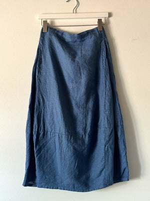 The CUT LOOSE - SIDE PLEAT SKIRT by CUT LOOSE hangs on a hanger against a plain grey background. The long white maxi skirt appears to be made of lightweight, slightly wrinkled linen fabric, with a simple design free of patterns or embellishments.