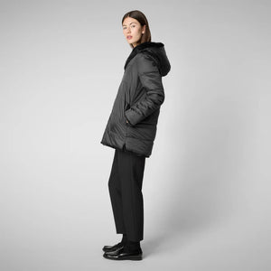 A person with straight dark hair is standing against a plain background, wearing the SAVE THE DUCK - BRIDGET REVERSIBLE FAUX FUR JACKET, black pants, and black shoes. The zipped-up coat by SAVE THE DUCK showcases sustainable fashion as the individual looks directly at the camera with a neutral expression.