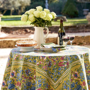 An outdoor round table set with a COULEUR NATURE JARDIN BLUE & VERT TABLECLOTH 71X71 displays a white pitcher with yellow roses, a pie, two red wine glasses, a bottle, and napkins amidst trees and shrubs in the garden.